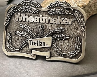 May include: A silver-toned belt buckle with the word "Wheatmaker" prominently displayed. The buckle features wheat stalk designs and the name "Treflan" on a banner. The overall design is rectangular with a textured background.
