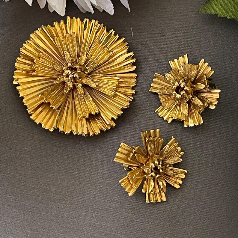 K&ouml;nnte beinhalten: Ein Set aus drei goldfarbenen Broschen in Form von stilisierten Blumen. Die gr&ouml;&szlig;te Brosche ist rund und die beiden kleineren Broschen sind sternf&ouml;rmig.