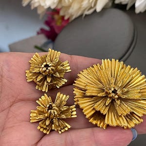 K&ouml;nnte beinhalten: Ein Set aus drei goldfarbenen Broschen in Blumenform. Die Broschen bestehen aus d&uuml;nnem, &uuml;bereinanderliegendem Metall, das einen zarten, strukturierten Look erzeugt.