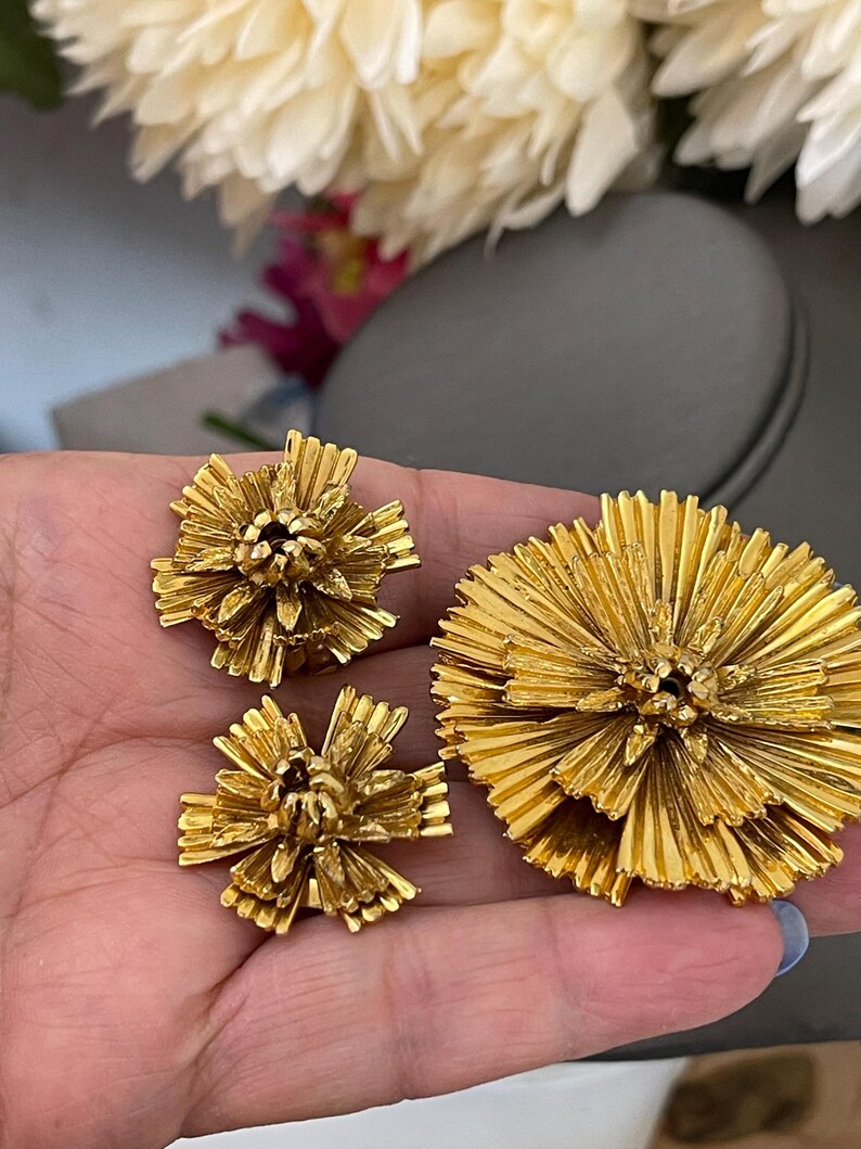 K&ouml;nnte beinhalten: Ein Set aus drei goldfarbenen Broschen in Blumenform. Die Broschen bestehen aus d&uuml;nnem, &uuml;bereinanderliegendem Metall, das einen zarten, strukturierten Look erzeugt.