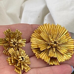 K&ouml;nnte beinhalten: Ein Set aus drei goldfarbenen Broschen in Blumenform. Die gr&ouml;&szlig;te Brosche befindet sich in der Mitte und die beiden kleineren Broschen befinden sich auf beiden Seiten. Die Broschen bestehen aus d&uuml;nnem, zartem Metall, das zu einer Blume mit vielen Bl&uuml;tenbl&auml;ttern geformt ist.