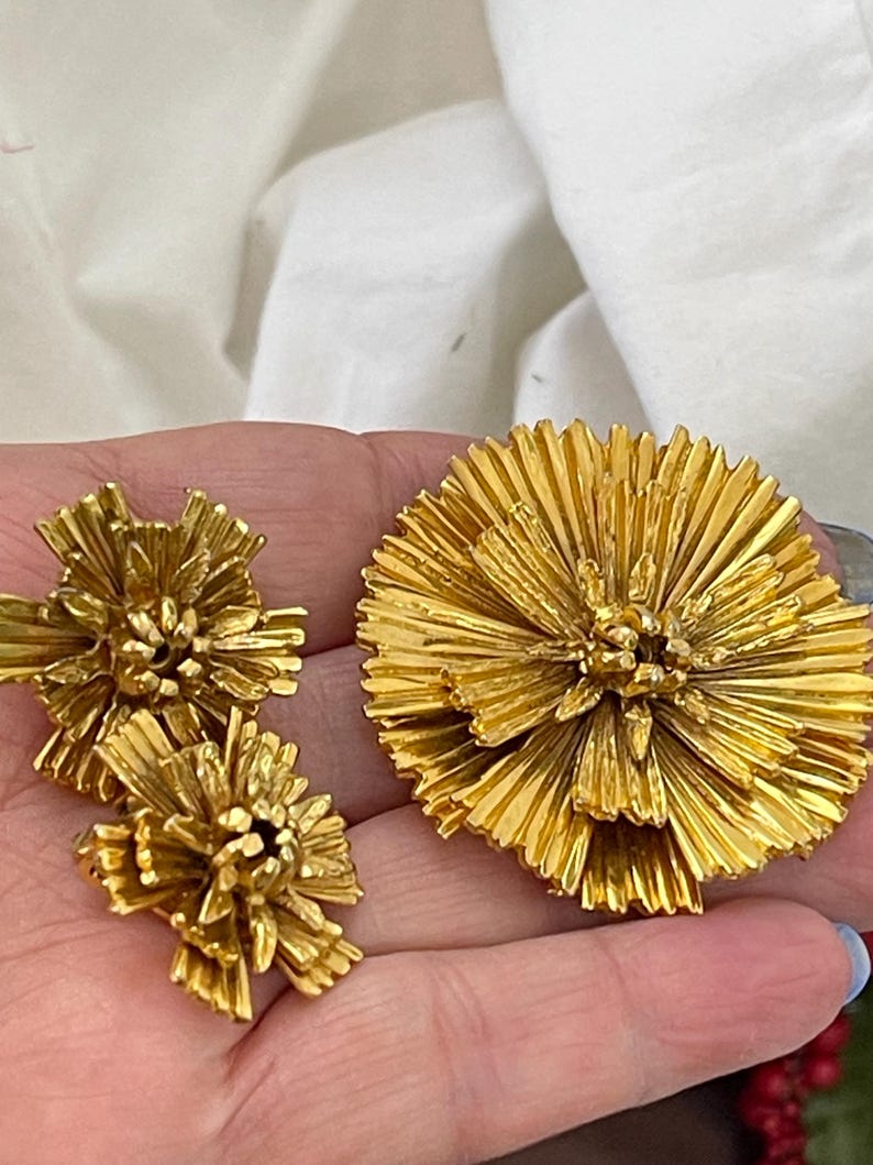 K&ouml;nnte beinhalten: Ein Set aus drei goldfarbenen Broschen in Blumenform. Die gr&ouml;&szlig;te Brosche befindet sich in der Mitte und die beiden kleineren Broschen befinden sich auf beiden Seiten. Die Broschen bestehen aus d&uuml;nnem, zartem Metall, das zu einer Blume mit vielen Bl&uuml;tenbl&auml;ttern geformt ist.