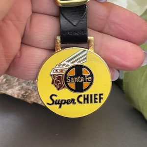 May include: A vintage Super Chief watch fob with a yellow face. The fob features the Santa Fe logo and the words "Super Chief" in black lettering. The fob is attached to a black leather strap with a gold-tone buckle.