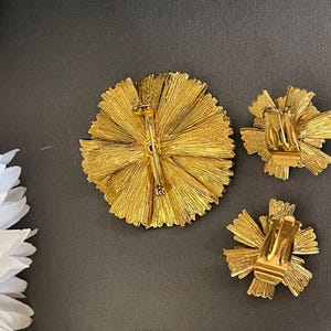 K&ouml;nnte beinhalten: Ein Set aus drei goldfarbenen Broschen in Form einer Blume. Die Broschen sind aus Metall und haben eine strukturierte Oberfl&auml;che. Die Broschen sind mit einem Bl&uuml;tenblatt-Cluster gestaltet, der sich von einem zentralen Punkt nach au&szlig;en erstreckt.