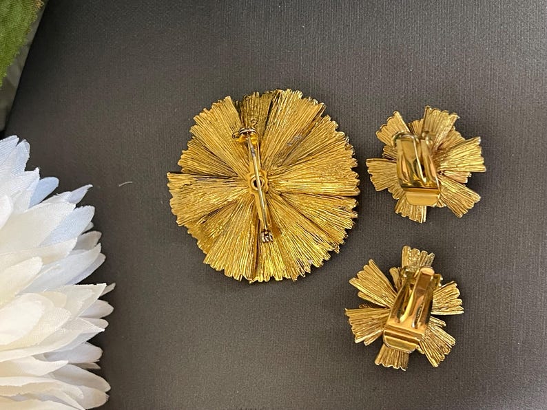 K&ouml;nnte beinhalten: Ein Set aus drei goldfarbenen Broschen in Form einer Blume. Die Broschen sind aus Metall und haben eine strukturierte Oberfl&auml;che. Die Broschen sind mit einem Bl&uuml;tenblatt-Cluster gestaltet, der sich von einem zentralen Punkt nach au&szlig;en erstreckt.