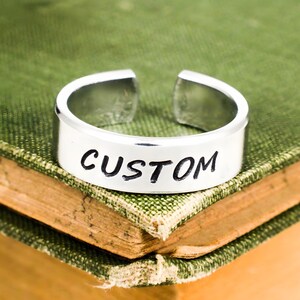 May include: A silver-colored metal ring with an open band design. The outer surface of the ring features the word CUSTOM engraved in black capital letters. The ring has a smooth, polished finish and a consistent width around the entire circumference.