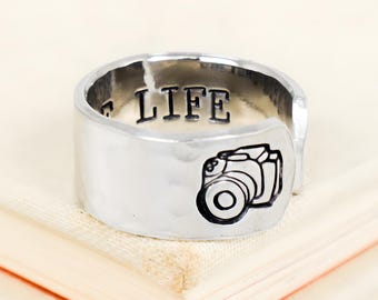 Pode incluir: Um anel de metal prateado com textura martelada, apresentando um ícone de câmara preto gravado na banda exterior e a palavra LIFE estampada em letras pretas na superfície interior da banda, que tem um design aberto.