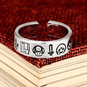 May include: A silver-toned open band ring featuring a series of engraved black icons around the outer surface, including a game cartridge, a mushroom, a sword, and a stylized creature, with a gap at the back for adjustable sizing.