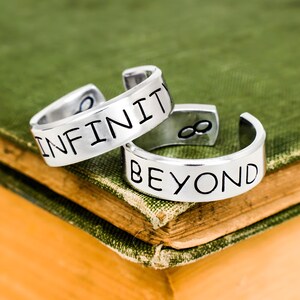 May include: Two silver-toned metal rings with an open-band design. One ring is stamped with the word INFINITY in black lettering, and the other ring is stamped with the word BEYOND in black lettering, accompanied by an infinity symbol icon.