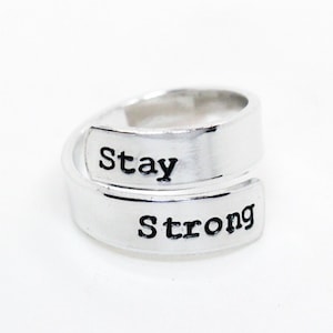 Puede incluir: Anillo plateado con las palabras "Stay Strong" grabadas. El anillo tiene un diseño único, con la banda envolviendo el dedo. El texto está en negrita, con una fuente sans-serif.