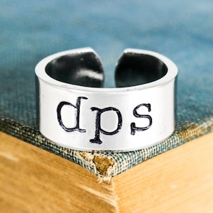 Peut inclure: Une bague en argent avec le mot "dps" gravé dessus. La bague est posée sur un livre bleu.