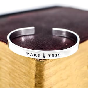 Pode incluir: Uma pulseira de metal cor de prata com design aberto na parte de trás. A frente da pulseira apresenta o texto preto estampado "TAKE" seguido por um pequeno ícone de espada e a palavra "THIS" em uma fonte sem serifa.