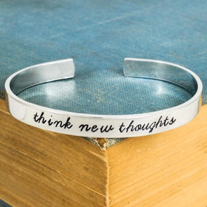 May include: Silver cuff bracelet with the inscription "think new thoughts" on a blue and brown book.