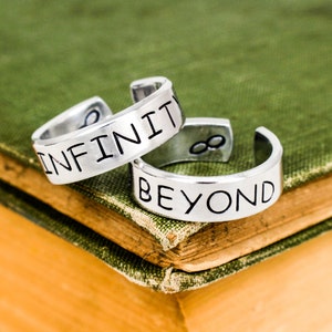May include: Two silver rings with the words "Infinity" and "Beyond" engraved on them. The rings are stacked on top of each other and are resting on a green book.