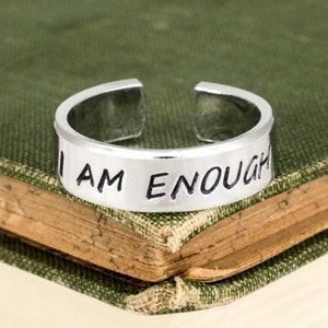 Op de afbeelding: Een zilveren ring met de woorden "I AM ENOUGH" gegraveerd erop. De ring ligt op een gesloten boek.