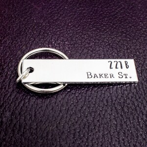 Pode incluir: Chaveiro de prata com uma etiqueta retangular que diz "221B Baker St."