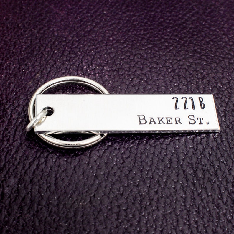 Pode incluir: Chaveiro de prata com uma etiqueta retangular que diz "221B Baker St."