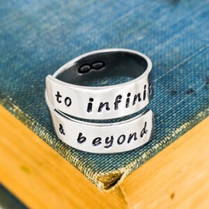 May include: Silver ring engraved with the words "to infinity & beyond". The ring is resting on a blue book.