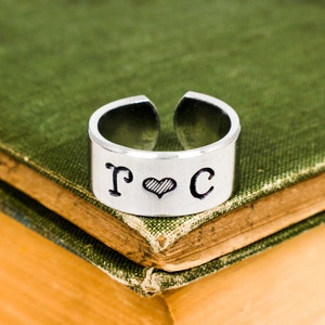 Puede incluir: Un anillo de plata con las letras "T" y "C" a cada lado de un pequeño corazón. El anillo está sobre un libro verde.