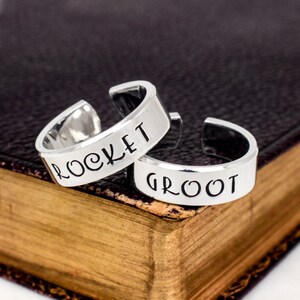May include: Two silver rings with the words "Rocket" and "Groot" engraved on them. The rings are on a brown leather-bound book.
