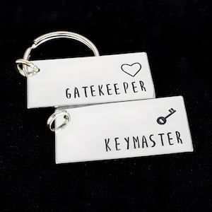 May include: Two rectangular silver-coloured metal keychains with split rings. One keychain is engraved with the word GATEKEEPER and a heart symbol. The other keychain is engraved with the word KEYMASTER and a small key icon in black lettering.