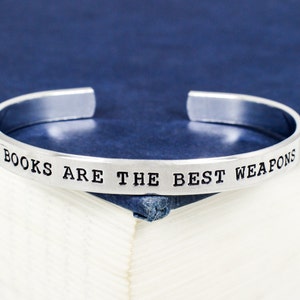 Op de afbeelding: Zilveren armband met de inscriptie "BOOKS ARE THE BEST WEAPONS" op een blauwe achtergrond.