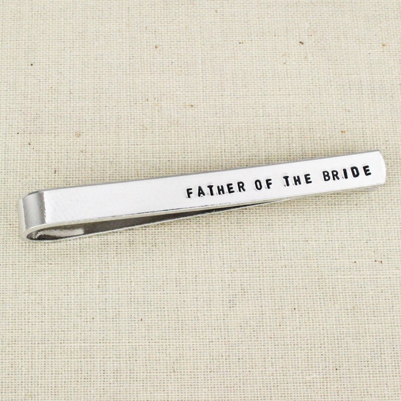 May include: A silver-colored tie bar with the text "FATHER OF THE BRIDE" in black, centered on the bar. The tie bar is rectangular with rounded edges and a curved clip on the back.