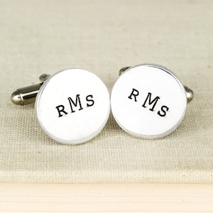 May include: A pair of silver cufflinks with the initials "RMS" engraved on each. The cufflinks are round and have a smooth, polished finish.