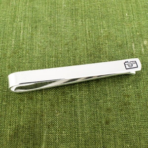 May include: Silver tie bar with a camera icon engraved on it. The tie bar is lying on a green fabric surface.