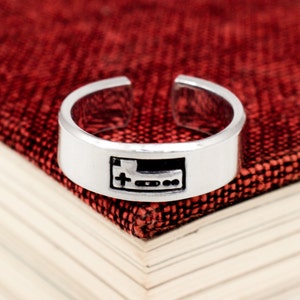 May include: A silver ring with a raised design of a video game controller. The ring is on a red fabric surface.