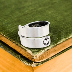May include: A silver ring with a heart design on the band. The ring is wrapped around a book with a green cover.
