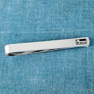 May include: A polished silver-tone metal tie bar featuring a small, black engraved graphic of a classic rectangular video game controller on the right end. The tie clip has a smooth, reflective surface and a simple, elongated rectangular shape.