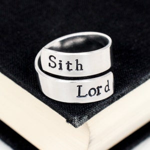 May include: A silver ring with the words "Sith Lord" engraved on it. The ring is wrapped around a book.