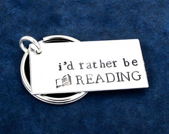 May include: A silver-toned rectangular metal keychain tag featuring the stamped text i'd rather be READING and a small icon of an open book. The tag is attached to a circular metal split ring, providing a functional accessory for keys.