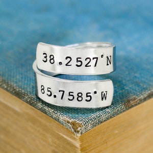 May include: A silver-colored wrap ring with stamped coordinates: 38.2527° N and 85.7585° W. The ring is displayed on a blue and tan book, showcasing its design and the contrast of the metal.