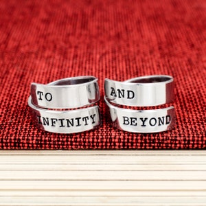 May include: Two silver rings with the words "To Infinity" and "And Beyond" engraved on them. The rings are on a red fabric background.