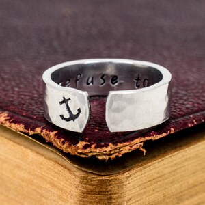 May include: A silver ring with a hammered finish and the words "refuse to" engraved on the band. An anchor symbol is engraved on the side of the ring.
