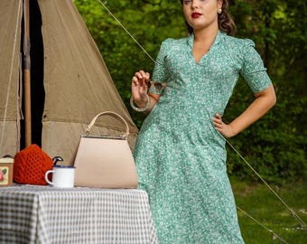 May include: A woman in a green floral dress poses near a vintage camping scene. A beige handbag, a picnic basket, and a small table with a checkered tablecloth are visible. A vintage lantern hangs from a tent.
