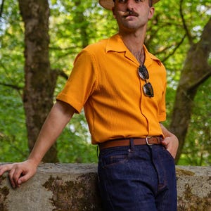 Puede incluir: Una persona viste una camisa naranja de manga corta, vaqueros azul oscuro y un sombrero de paja con una banda a rayas. El atuendo incluye un cinturón marrón y gafas de sol. El fondo presenta árboles verdes.