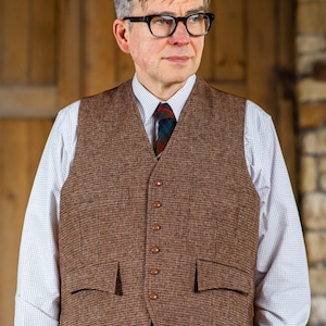 Puede incluir: Un chaleco de tweed marr&oacute;n con un traje a juego. El chaleco tiene una parte delantera abotonada y dos bolsillos delanteros. La persona lleva una camisa blanca con un estampado de cuadros azules y blancos y una corbata estampada. La persona tambi&eacute;n lleva gafas negras.