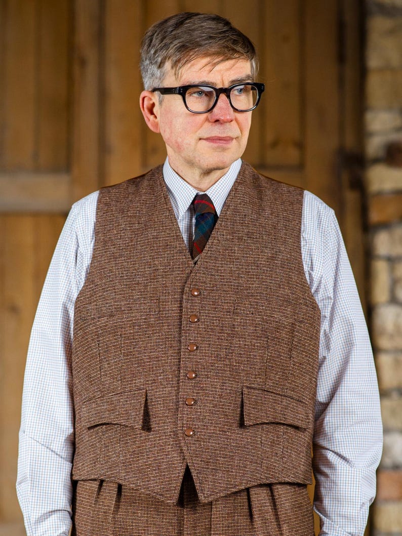 Puede incluir: Un chaleco de tweed marr&oacute;n con un traje a juego. El chaleco tiene una parte delantera abotonada y dos bolsillos delanteros. La persona lleva una camisa blanca con un estampado de cuadros azules y blancos y una corbata estampada. La persona tambi&eacute;n lleva gafas negras.
