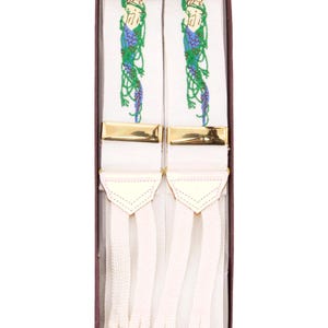 May include: White suspenders with gold-colored clasps and decorative embroidery. The embroidery features a woman's head and flowing vines in green and purple. The suspenders are presented in a brown box.