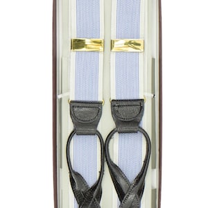 Può includere: Bretelle a righe blu e bianche con estremità in pelle nera e chiusure metalliche color oro, presentate in una scatola marrone. Le bretelle hanno un design classico, adatto per abbigliamento formale o business.
