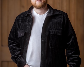 May include: A man with a beard and glasses is wearing a black corduroy shirt over a white t-shirt and dark blue jeans.