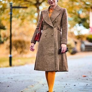 Puede incluir: Una mujer con un abrigo de lana marrón de doble botonadura, medias naranjas y zapatos marrones. También lleva gafas y sostiene un bolso rojo y guantes marrones. El fondo es un parque borroso con colores otoñales.
