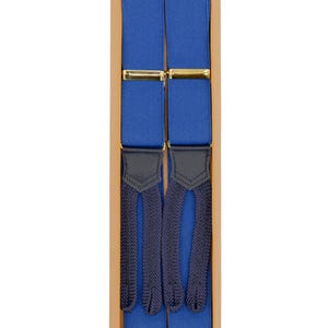 May include: A pair of blue suspenders with gold-colored clasps and dark blue leather accents, presented in a cardboard box. The suspenders are a vibrant blue color, with dark blue woven ends.