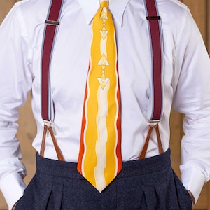 May include: A crisp white button-down shirt paired with a bright yellow necktie featuring orange side panels and a central white geometric pattern. The outfit includes deep burgundy braces with light blue trim and brown leather attachments.