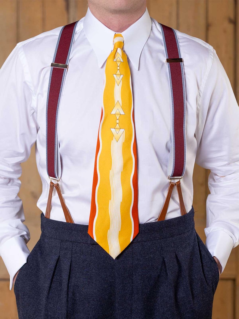 May include: A crisp white button-down shirt paired with a bright yellow necktie featuring orange side panels and a central white geometric pattern. The outfit includes deep burgundy braces with light blue trim and brown leather attachments.