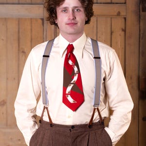 May include: A cream-coloured button-down shirt with a red tie featuring a brown and white design. The outfit includes brown trousers and grey braces with leather attachments. The person is standing in front of a wooden door.