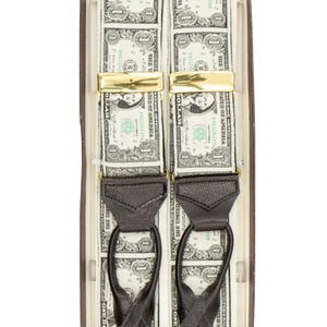 May include: A pair of novelty suspenders featuring a pattern of US one-dollar bills. The suspenders have gold-colored clasps and black leather accents. They are presented in a brown box.